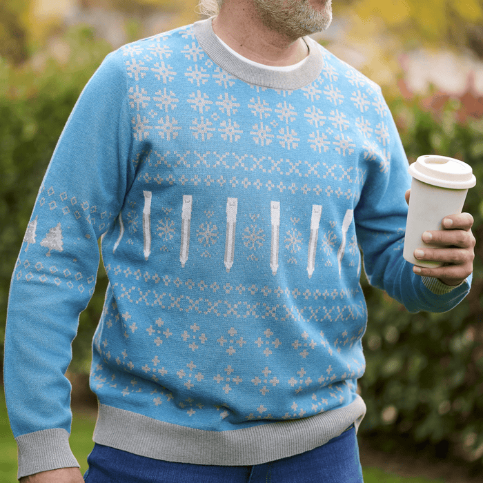 Man holding a coffee and wearing a blue and grey holiday sweater featuring pencils and snowflakes