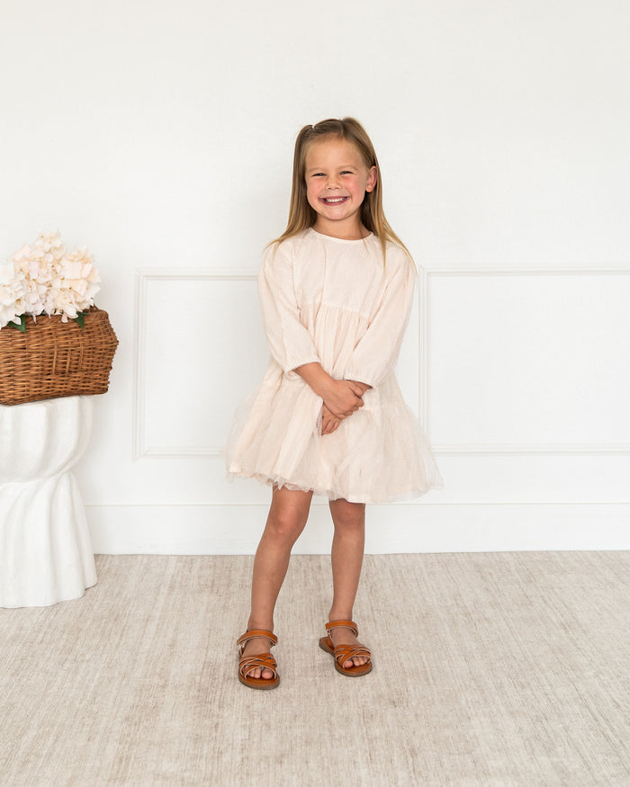 A toddler young girl with blonde hair smiling and wearing a light beige long-sleeve organic tulle dress and brown leather sandals, standing in a bright room with a white wall and light beige textured floor.