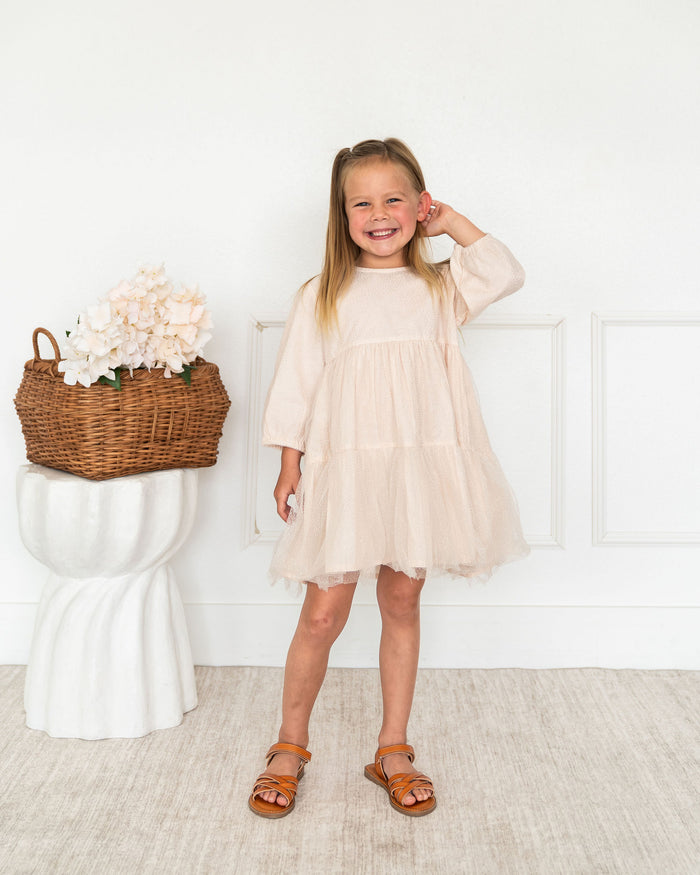 A young girl with blonde hair stands smiling in a room, wearing a light beige tiered tulle dress with three-quarter sleeves and brown sandals. To her left is a white pedestal holding a wicker basket filled with pale pink flowers.