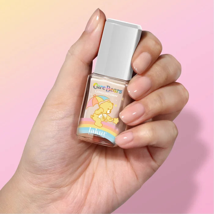 A hand holds a bottle of LONDONTOWN Good Vibes Only nail polish featuring a joyful Care Bears design, set against a warm sunlit peach and yellow gradient background.