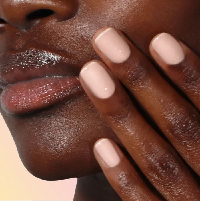 Close-up of a person's face and hand with sunlit peach, glossy nude-colored Good Vibes Only nail polish by LONDONTOWN and natural, shiny lips, creating a warm, inviting vibe.