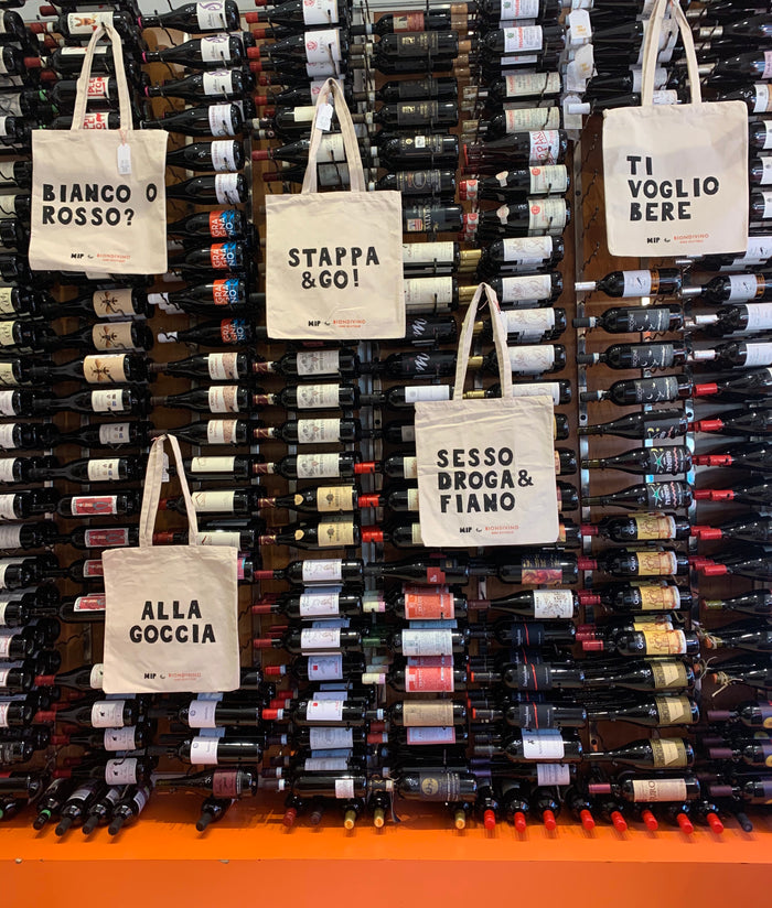 Canvas tote bags with printed phrases, loop handles, displayed among wine bottles
